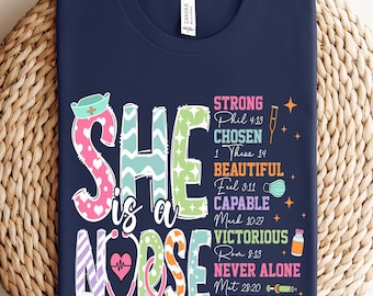 May include: A navy blue t-shirt with a colorful graphic that says "She is a Nurse" with a stethoscope and other medical symbols. The graphic also includes bible verses about being strong, chosen, beautiful, capable, victorious, never alone, and greatly loved.