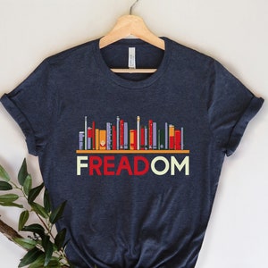 May include: A dark blue t-shirt with a graphic of a bookshelf full of colorful books. The text "FREEDOM" is printed below the bookshelf.