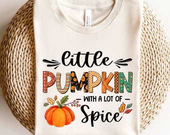 May include: A white t-shirt with the text "little pumpkin with a lot of spice" in black and orange. The word "pumpkin" is made up of colorful patchwork pieces. A small orange pumpkin with green leaves is in the center of the text.