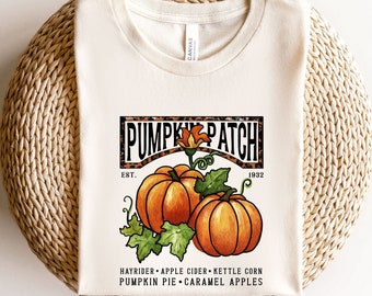 May include: Cream-colored t-shirt featuring a "PUMPKIN PATCH" design with two orange pumpkins, green leaves, and the text "EST. 1932." Additional text includes "HAYRIDER • APPLE CIDER • KETTLE CORN • PUMPKIN PIE • CARAMEL APPLES."