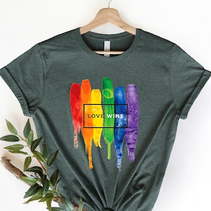 May include: A dark green t-shirt with a rainbow paint splatter design and the text "LOVE WINS" in a black box.