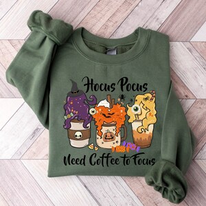 May include: Green sweatshirt with a Halloween-themed design featuring three coffee cups with spooky faces and the text "Hocus Pocus Need Coffee to Focus".