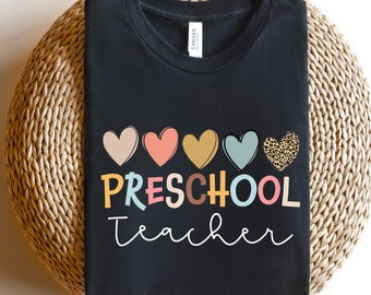May include: Black t-shirt with the text "PRESCHOOL teacher" in colorful letters and a row of colorful hearts above the text. One heart has a leopard print pattern.