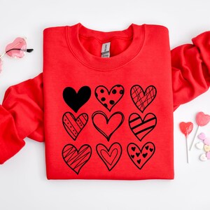 May include: Red sweatshirt with a graphic of nine black hearts with different patterns on the front. The hearts are arranged in a grid pattern.