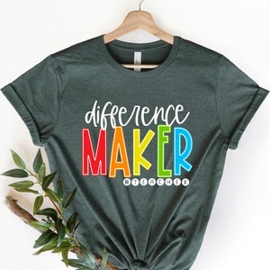 May include: A dark green t-shirt with the text "difference maker" in rainbow colors. The text "#teacher" is below the main text in smaller white letters.