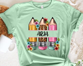 May include: A light green t-shirt with a graphic of three pencils with floral accents and the name "ARJA" printed on a white banner.