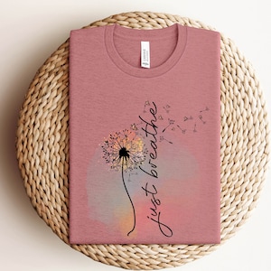 May include: A dusky pink t-shirt with a dandelion design and the words "just breathe" in cursive. The dandelion is black and white, set against a watercolour background of pink, orange, and grey. The shirt is displayed on a woven wicker mat.