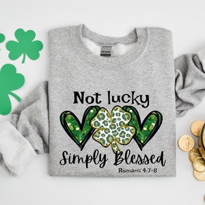 Puede incluir: Sudadera gris con un tr&eacute;bol verde y dos corazones con un dise&ntilde;o de purpurina dorada. El texto dice "Not lucky Simply Blessed Romans 4:7-8".