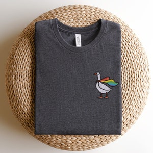 May include: A dark grey t-shirt with a white goose wearing a rainbow cape. The goose is facing right and has a small black beak. The t-shirt has a white tag that says "Canvas" on it.