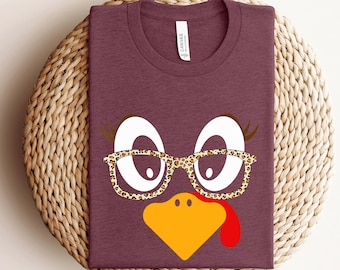 Puede incluir: Una camiseta color marrón con un gráfico de cara de pavo. El pavo tiene ojos blancos, un pico amarillo y una papada roja. El pavo lleva gafas de estampado de leopardo.