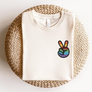 K&ouml;nnte beinhalten: Ein wei&szlig;es T-Shirt mit einem Regenbogen-Friedenszeichen-Motiv. Das Shirt ist gefaltet und auf einer gewebten Matte ausgestellt.