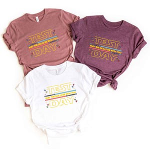 May include: Three t-shirts in shades of mauve, purple, and white. Each shirt features the text "TEST DAY" in yellow, with the phrase "MAY THE SKILLS BE WITH YOU" below. The design includes colorful lines and star-like accents.