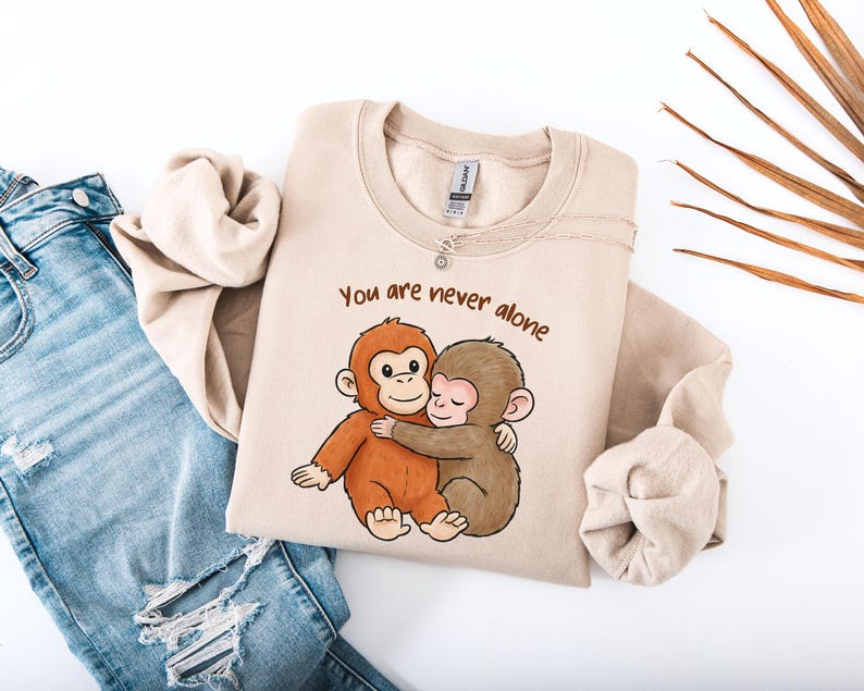 Puede incluir: Sudadera beige con el texto "You are never alone" y una ilustraci&oacute;n de dibujos animados de dos monos abraz&aacute;ndose. La sudadera est&aacute; doblada junto a unos vaqueros azules.