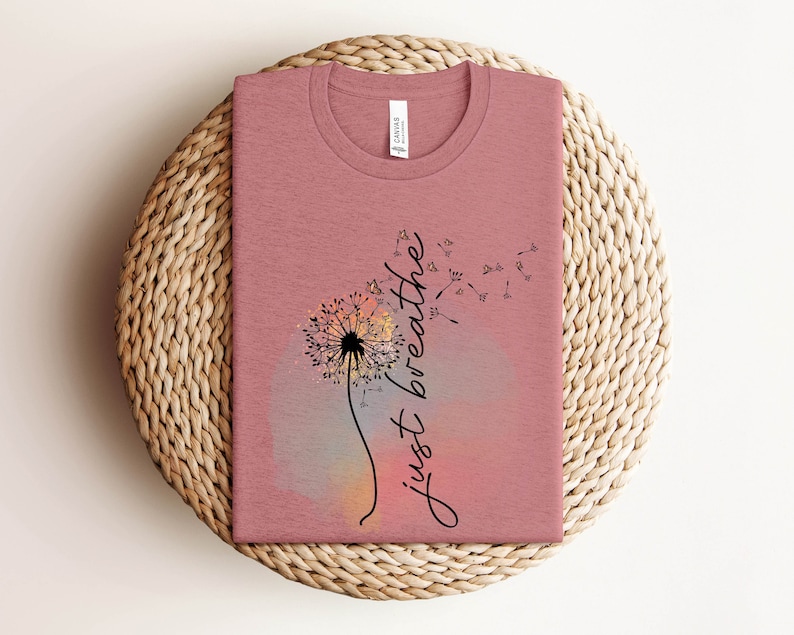 May include: A dusky pink t-shirt with a dandelion design and the words "just breathe" in cursive. The dandelion is black and white, set against a watercolour background of pink, orange, and grey. The shirt is displayed on a woven wicker mat.