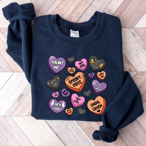 Puede incluir: Una sudadera azul marino con un dise&ntilde;o de tem&aacute;tica de Halloween con corazones de caramelo con frases como "Haunt Me", "Spooky Vibes" y "Ghoul Crush".