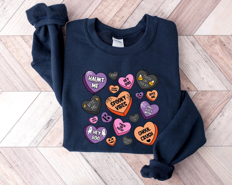 Puede incluir: Una sudadera azul marino con un dise&ntilde;o de tem&aacute;tica de Halloween con corazones de caramelo con frases como "Haunt Me", "Spooky Vibes" y "Ghoul Crush".