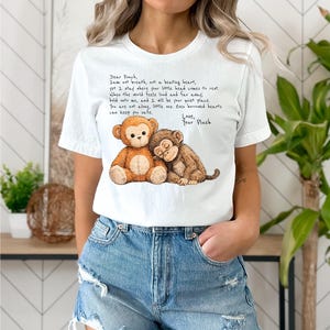 K&ouml;nnte beinhalten: Wei&szlig;es T-Shirt mit einer herzerw&auml;rmenden Illustration von zwei Stoffaffen. Einer ist hellbraun, der andere dunkelbraun, und sie umarmen sich. Das Shirt hat auch einen aufgedruckten Text.