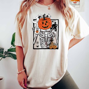K&ouml;nnte beinhalten: Ein wei&szlig;es T-Shirt mit einem Aufdruck einer K&uuml;rbis mit einem Skelettk&ouml;rper, der eine Tasse mit der Aufschrift "Happy Halloween" h&auml;lt.