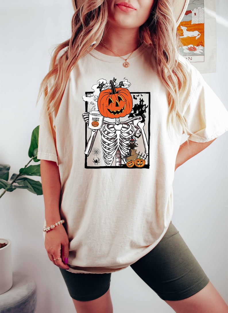 K&ouml;nnte beinhalten: Ein wei&szlig;es T-Shirt mit einem Aufdruck einer K&uuml;rbis mit einem Skelettk&ouml;rper, der eine Tasse mit der Aufschrift "Happy Halloween" h&auml;lt.