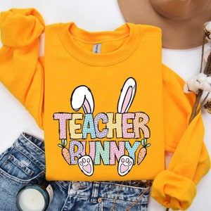 May include: Yellow sweatshirt with a graphic of a bunny with the text "Teacher Bunny" in rainbow colors. The bunny has pink ears and paws.
