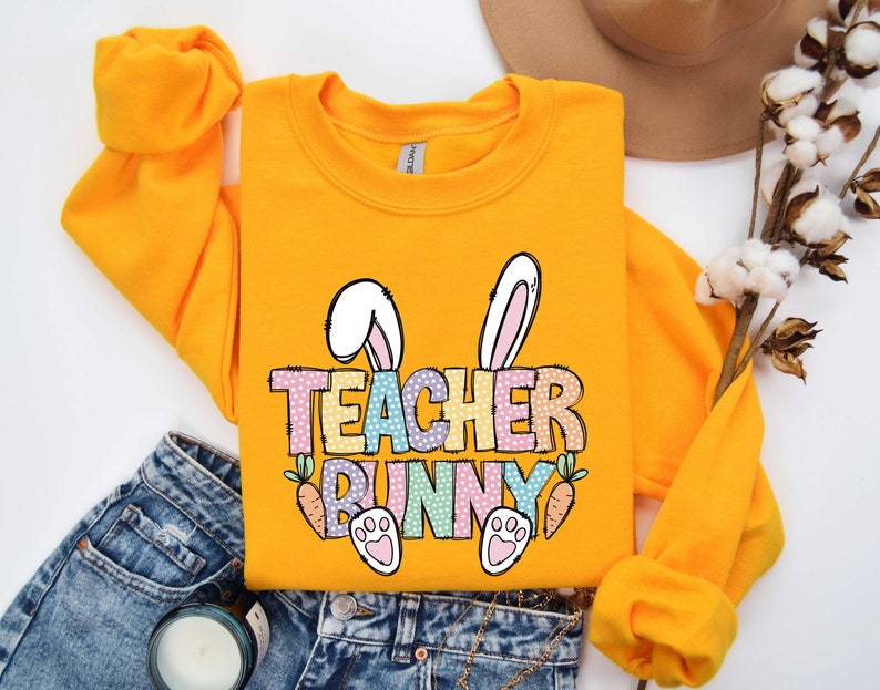 May include: Yellow sweatshirt with a graphic of a bunny with the text "Teacher Bunny" in rainbow colors. The bunny has pink ears and paws.
