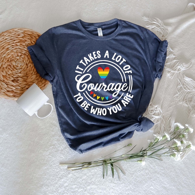 May include: Navy blue t-shirt with the text "It takes a lot of Courage to be who you are" in white, with a rainbow heart and rainbow accents. The shirt is folded and tied at the bottom.