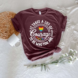 May include: Burgundy t-shirt with the text "It Takes a Lot of Courage to Be Who You Are" in a white circular design. The design includes a rainbow heart and rainbow colored triangles. The shirt is knotted at the waist.
