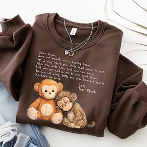 K&ouml;nnte beinhalten: Ein braunes Sweatshirt mit einer Grafik von zwei Affen und dem Text "Dear Punch, I am not breath, not a beating heart..." Eine silberne Halskette liegt &uuml;ber dem Sweatshirt. Das Sweatshirt hat einen Rundhalsausschnitt und lange &Auml;rmel.