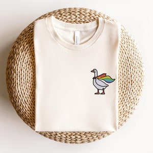 May include: A white t-shirt with a cartoon goose wearing a rainbow cape. The goose is facing right and has a black outline. The t-shirt is on a woven straw surface.