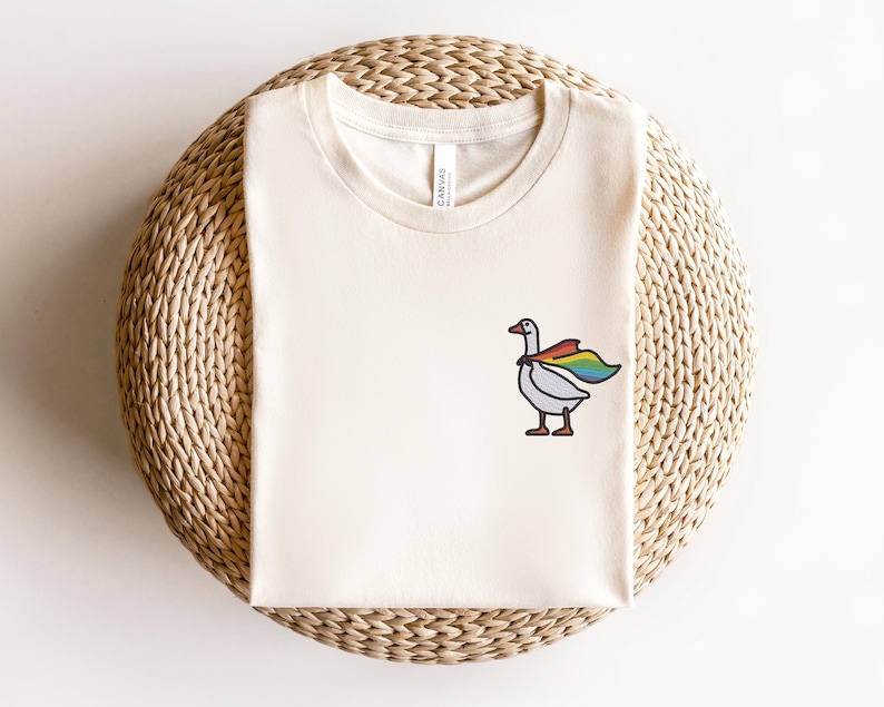May include: A white t-shirt with a cartoon goose wearing a rainbow cape. The goose is facing right and has a black outline. The t-shirt is on a woven straw surface.