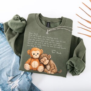 K&ouml;nnte beinhalten: Olivgr&uuml;nes Sweatshirt mit einer Grafik von zwei Cartoon-Affen und dem Text "Dear Peach". Eine silberne Halskette ist &uuml;ber dem Sweatshirt drapiert. Das Sweatshirt liegt neben einer Jeans.