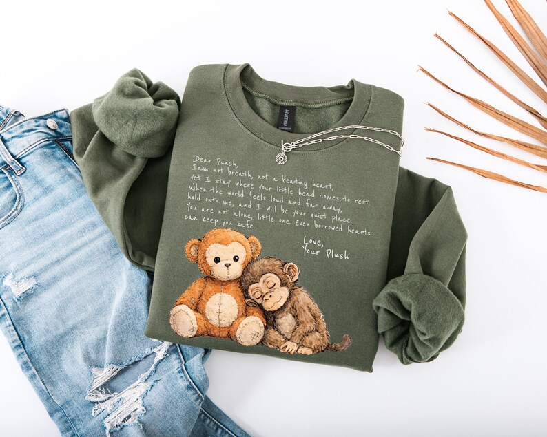 K&ouml;nnte beinhalten: Olivgr&uuml;nes Sweatshirt mit einer Grafik von zwei Cartoon-Affen und dem Text "Dear Peach". Eine silberne Halskette ist &uuml;ber dem Sweatshirt drapiert. Das Sweatshirt liegt neben einer Jeans.