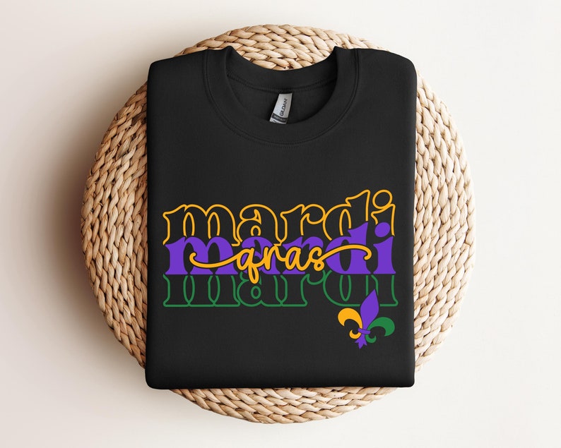 May include: A black t-shirt with the text 'Mardi Gras' in yellow, green, and purple on it. The text is in a layered style with the word 'Mardi' on top of the word 'gras' and the word 'Mardi' on top of the word 'gras'. There is a purple and gold fleur-de-lis below the text.