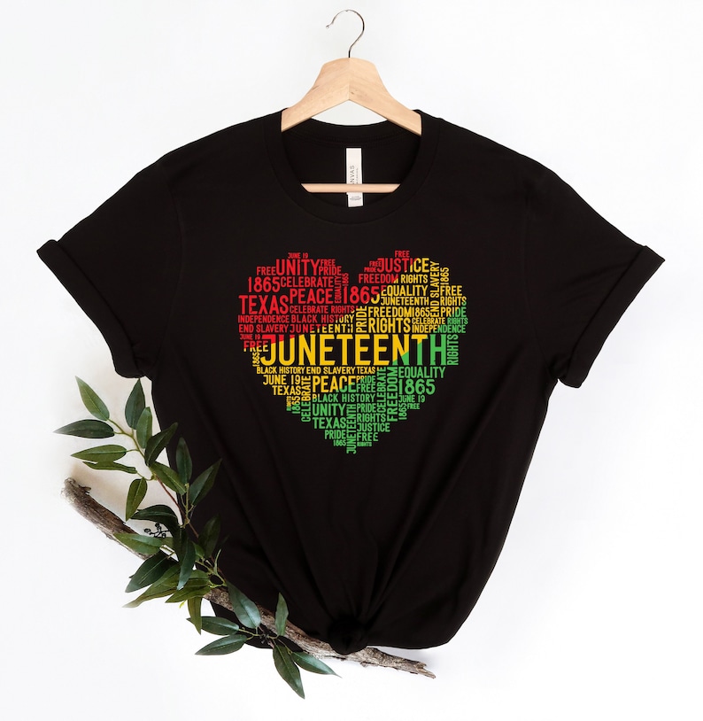 May include: Black t-shirt with a heart-shaped design composed of words in red, yellow, and green. The words include "Juneteenth," "Texas," "Freedom," and "Rights." The shirt is hanging on a wooden hanger.