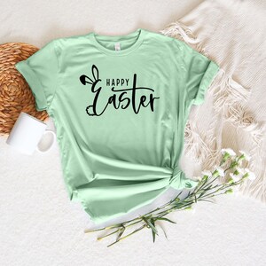 Puede incluir: Una camiseta verde claro con un dise&ntilde;o de orejas de conejo negras y el texto "Happy Easter" en letra cursiva.