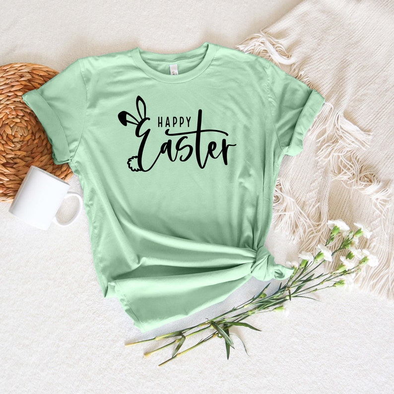 Puede incluir: Una camiseta verde claro con un dise&ntilde;o de orejas de conejo negras y el texto "Happy Easter" en letra cursiva.