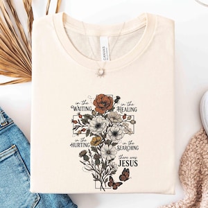 May include: Cream-colored t-shirt with a floral cross design. The cross is composed of various flowers in shades of orange, white, and yellow, with two butterflies. Text on the shirt reads "in the waiting, in the healing, in the hurting, in the searching, there was Jesus."