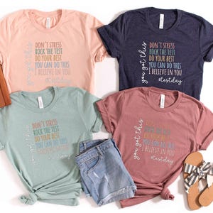 May include: Four t-shirts in various colors: peach, navy, dusty rose, and seafoam green. Each shirt has the text "Don't Stress, Rock the Test, Do Your Best, You Can Do This, I Believe in You" and "You Got This" printed on them. Accessories include denim shorts, sandals, and a brown clutch.