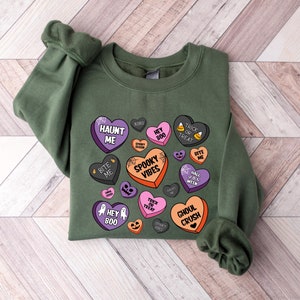 Puede incluir: Sudadera verde con un dise&ntilde;o de tem&aacute;tica de Halloween con corazones de caramelo con frases como "Haunt Me", "Spooky Vibes" y "Ghoul Crush".