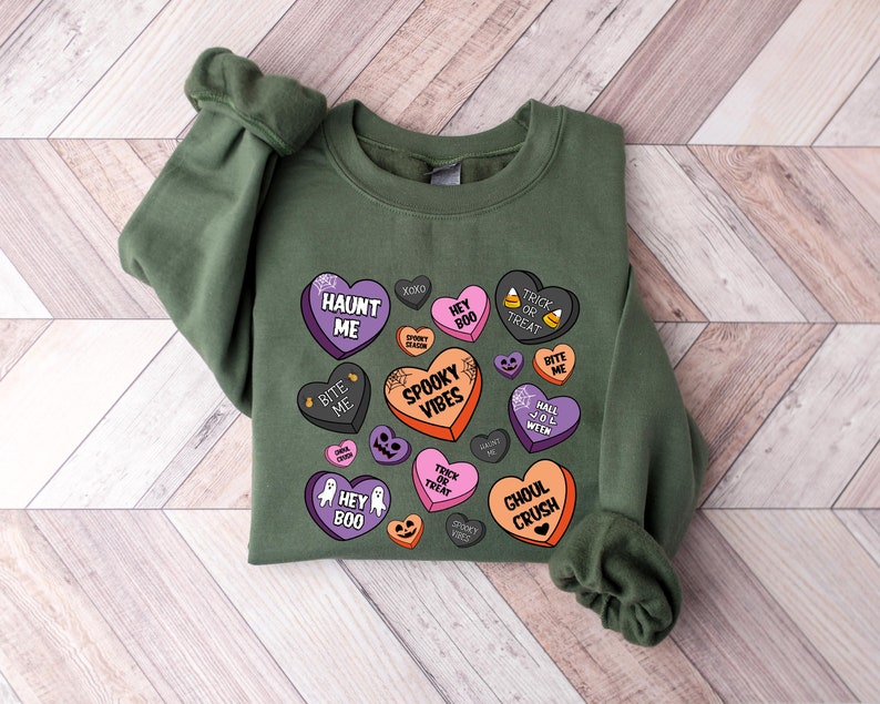 Puede incluir: Sudadera verde con un dise&ntilde;o de tem&aacute;tica de Halloween con corazones de caramelo con frases como "Haunt Me", "Spooky Vibes" y "Ghoul Crush".