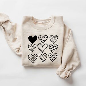 May include: A beige sweatshirt featuring a black graphic of nine different hearts. The hearts are all in different styles and have various patterns.