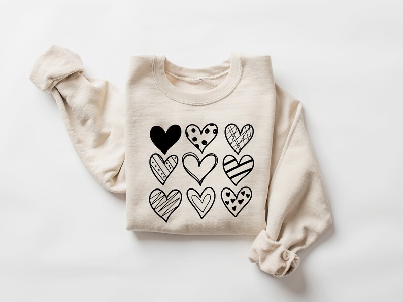 May include: A beige sweatshirt featuring a black graphic of nine different hearts. The hearts are all in different styles and have various patterns.