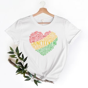 May include: White t-shirt with a heart-shaped design filled with words in red, yellow, and green. The words include "Juneteenth," "Texas," "Freedom," and "Equality." The shirt is hanging on a wooden hanger.