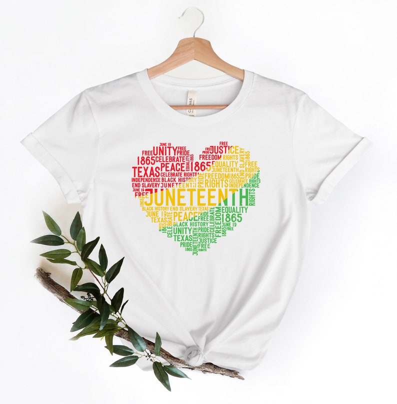 May include: White t-shirt with a heart-shaped design filled with words in red, yellow, and green. The words include "Juneteenth," "Texas," "Freedom," and "Equality." The shirt is hanging on a wooden hanger.