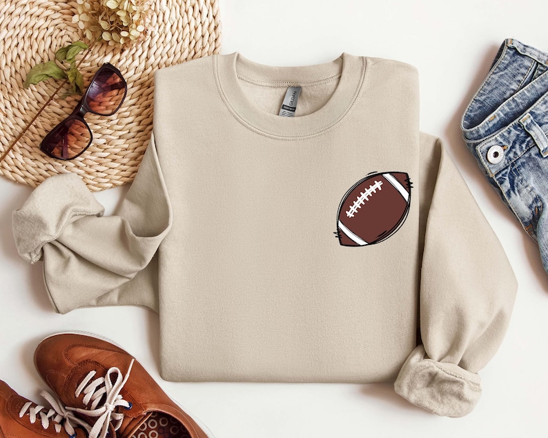 May include: A tan sweatshirt with a brown and white football design on the front.