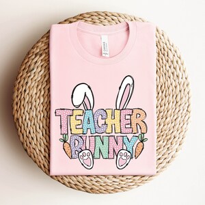 May include: Pink t-shirt with a graphic of a bunny with the text "Teacher Bunny" in rainbow colors with white polka dots.