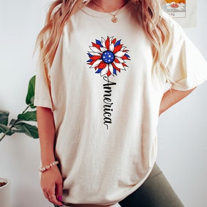 Puede incluir: Una camiseta blanca con un dise&ntilde;o de girasol rojo, blanco y azul y la palabra "America" escrita verticalmente debajo de la flor.