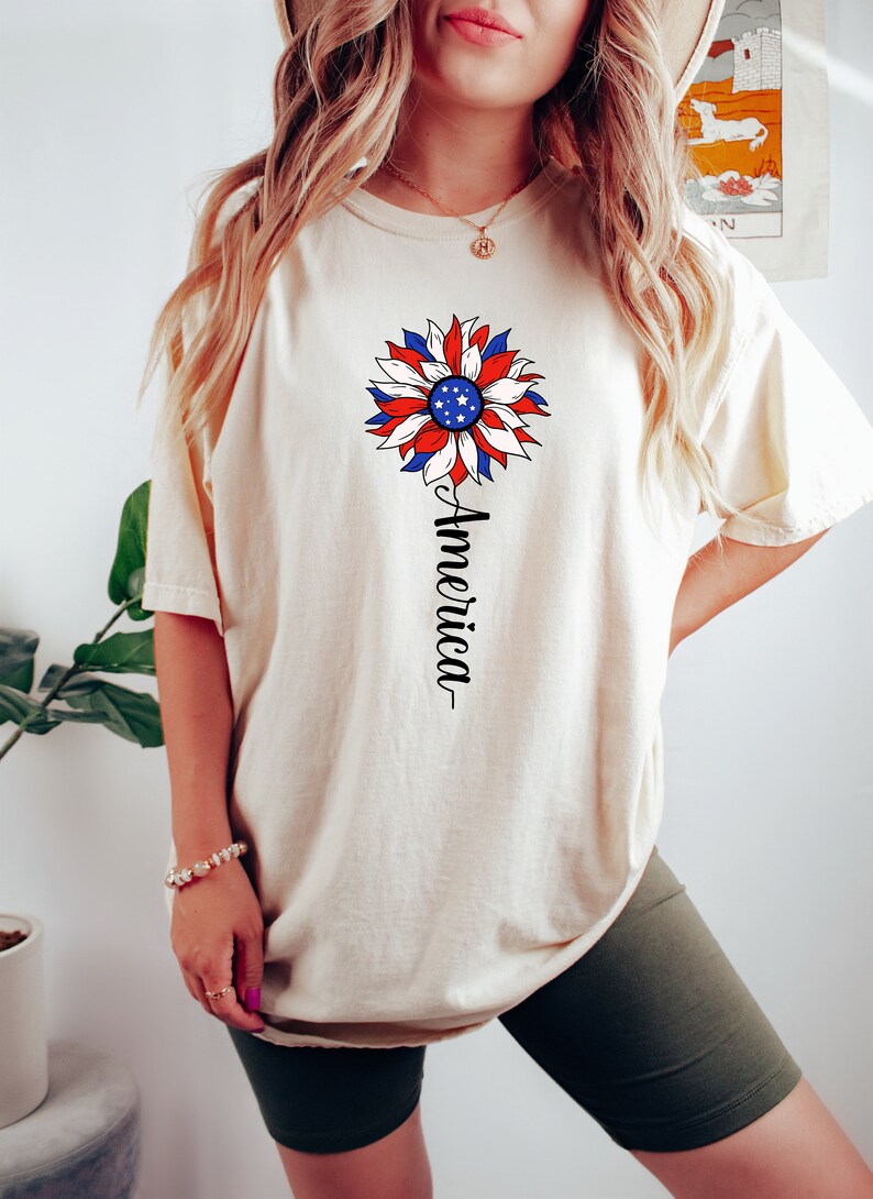 Puede incluir: Una camiseta blanca con un dise&ntilde;o de girasol rojo, blanco y azul y la palabra "America" escrita verticalmente debajo de la flor.