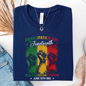 May include: Navy blue t-shirt with the words "Emancipation Day Juneteenth" in green and white script. The shirt features three raised fists in green, yellow, and red, with the text "Black American Freedom" and "June 19th 1865". A silver necklace is on the shirt.