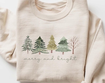 May include: A cream-colored sweatshirt featuring a watercolor design of five Christmas trees in varying shades of green and gold. The phrase "merry and bright" is written in a delicate script below the trees.